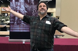 Image of a man, Chuck Lukacs, standing with outspread arms in front of a painting he created.