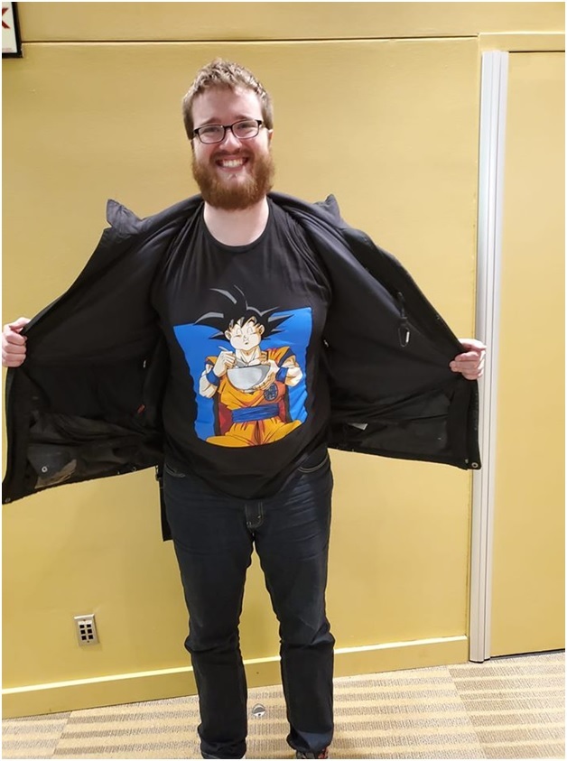 Image of a white, bearded man holding open his jacket to display a t-shirt of Goku from the Dragonball franchise eating from a bowl.